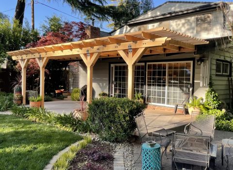 A wooden shade structure at the back of a house next to a garden